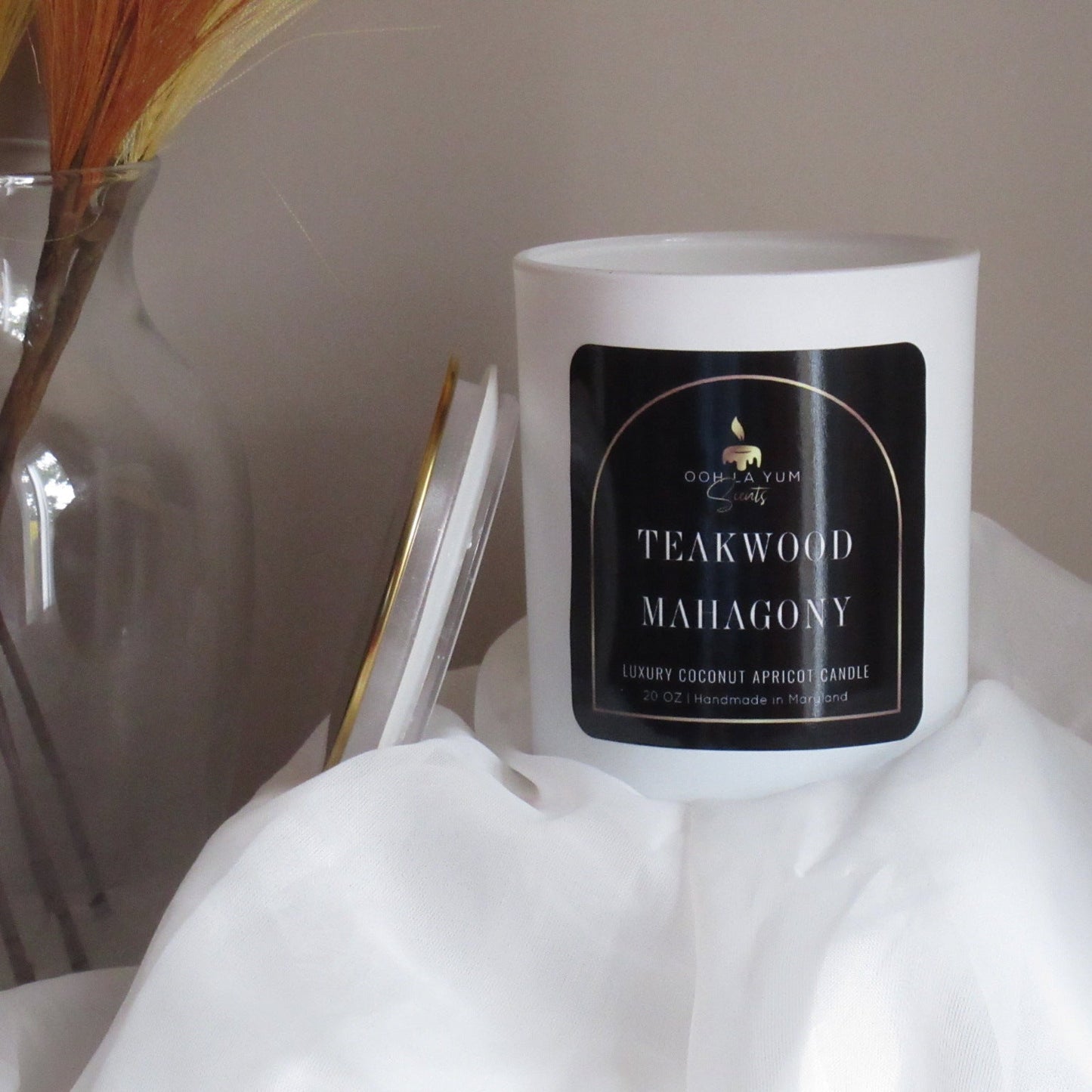 Candle with a label on a white surface, with a vase and decorative items in the background.