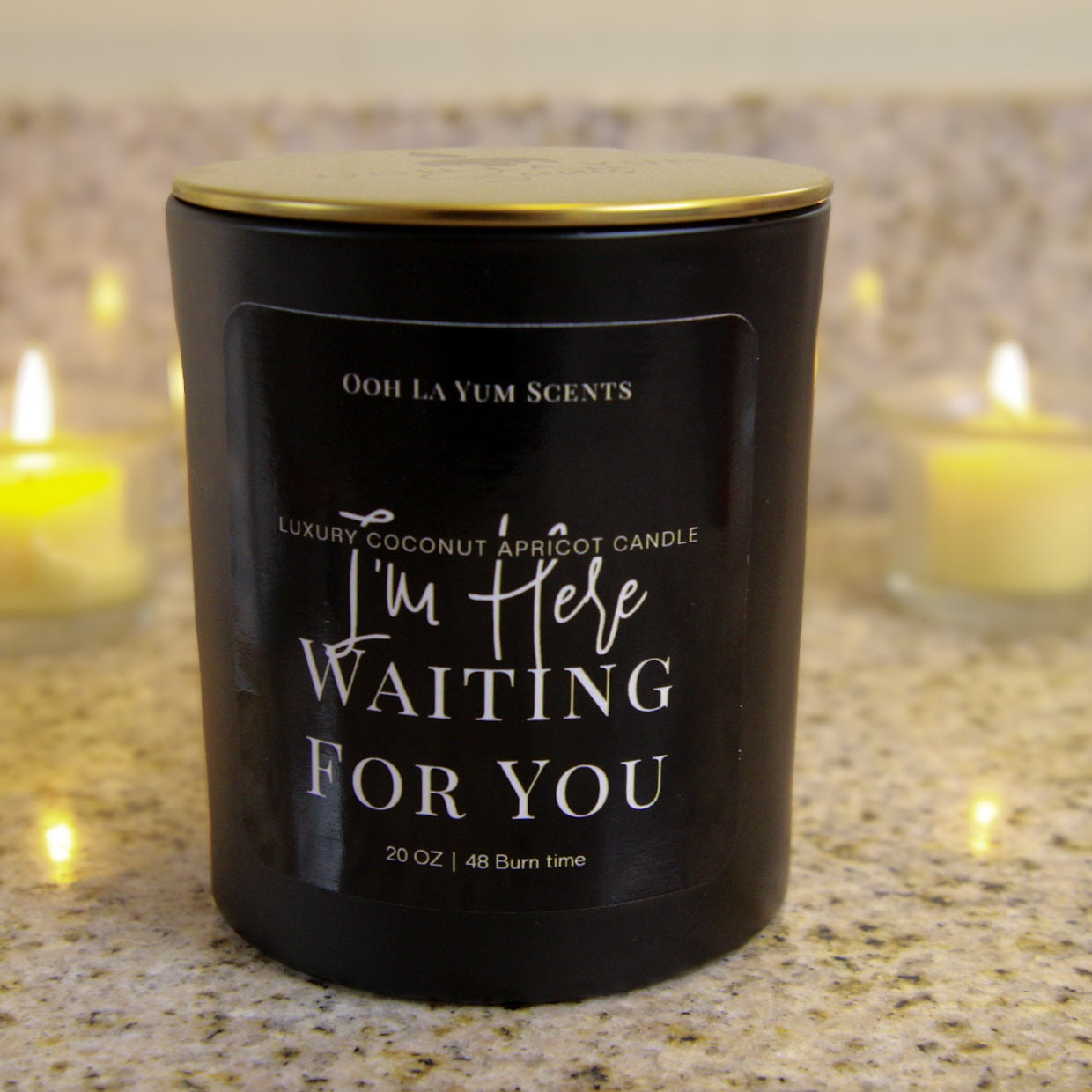 Black candle with gold lid labeled 'I'm Here Waiting For You' by Ooh La Yum Scents on a countertop.