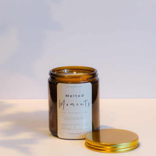 Candle in an amber glass jar with a gold lid on a light background with label 'melted moments'